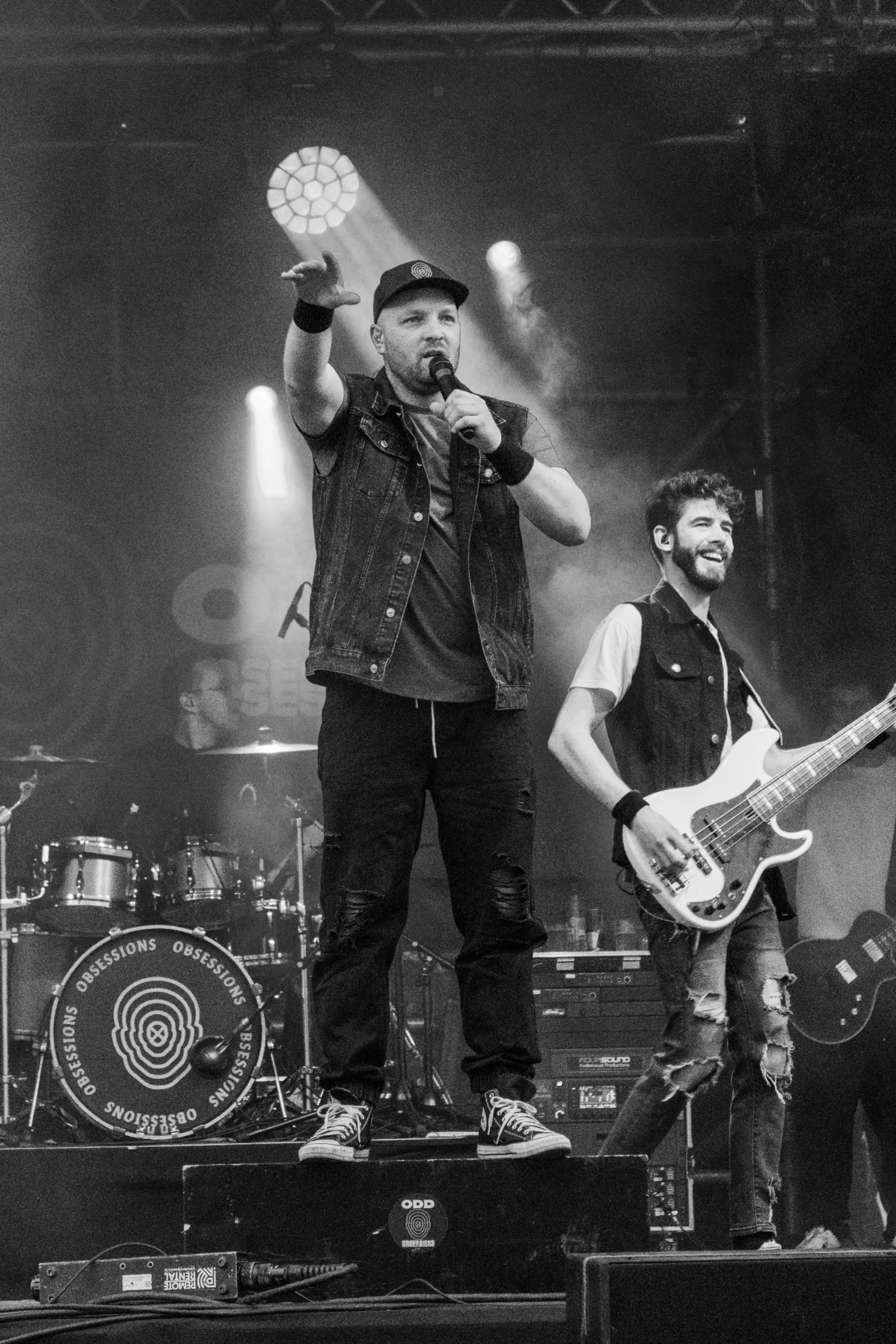 Zwart-wit shot van zanger met cap en spijkergilet op monitor met opgeheven arm naast lachende bassist met witte basgitaar