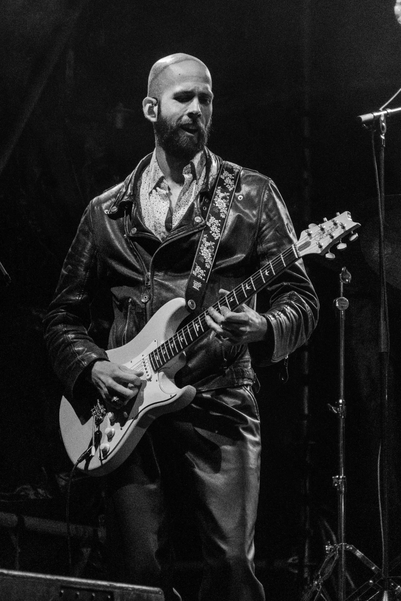 Zwart-wit portret van gitarist met baard en leren jack die PRS gitaar bespeelt op podium