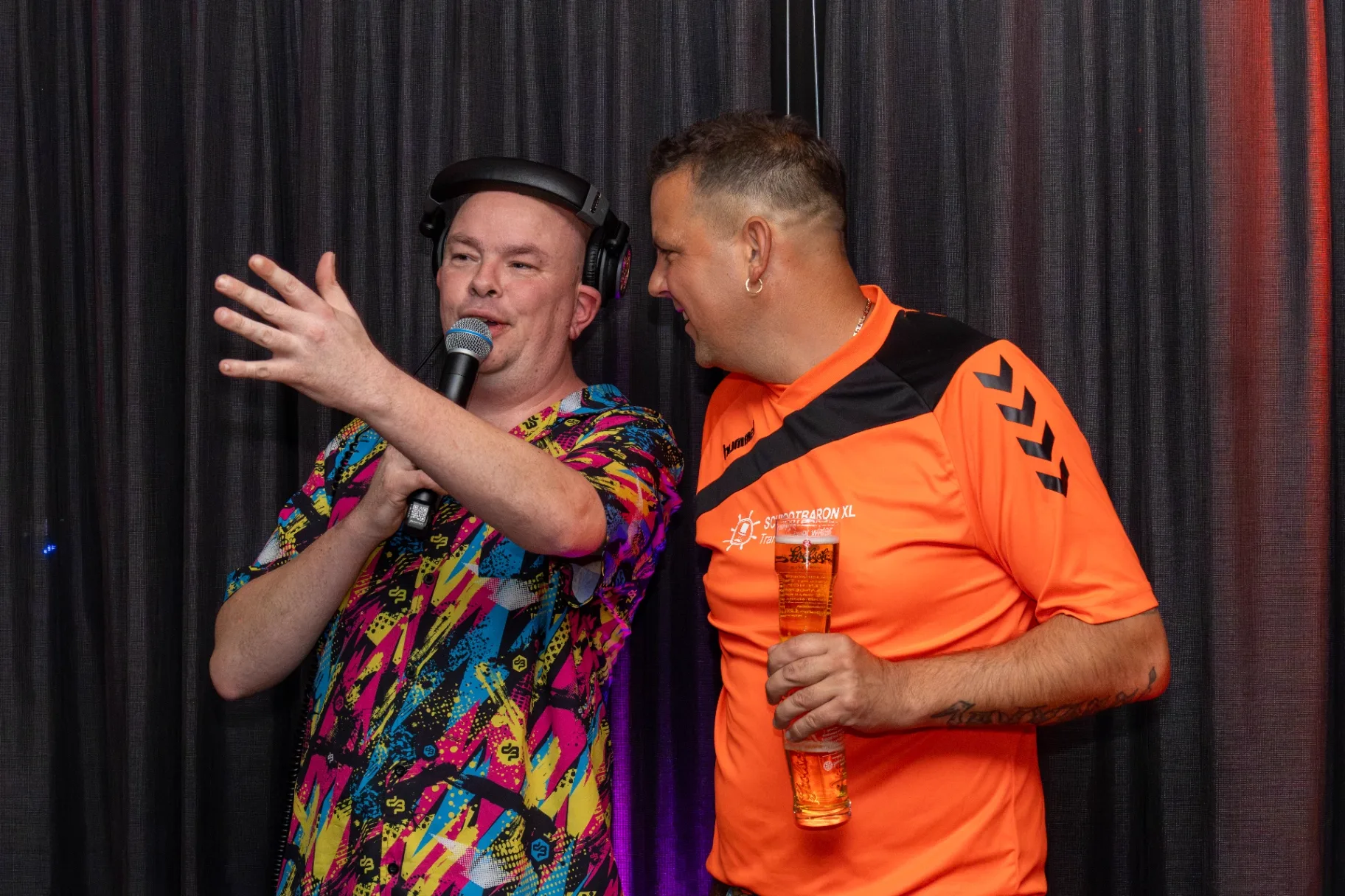 Zanger met microfoon en gast in oranje shirt zingen samen op feest