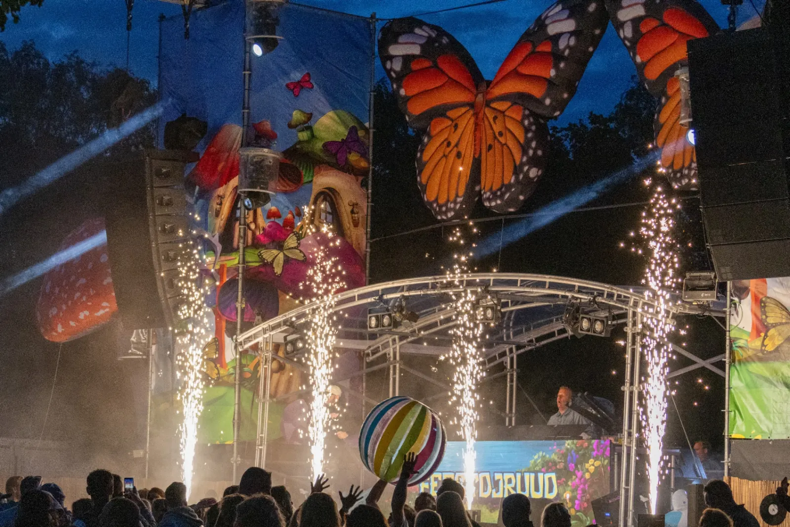 Festivalpodium met vuurwerk fonteinen, vlinderdecors en juichend publiek bij schemering