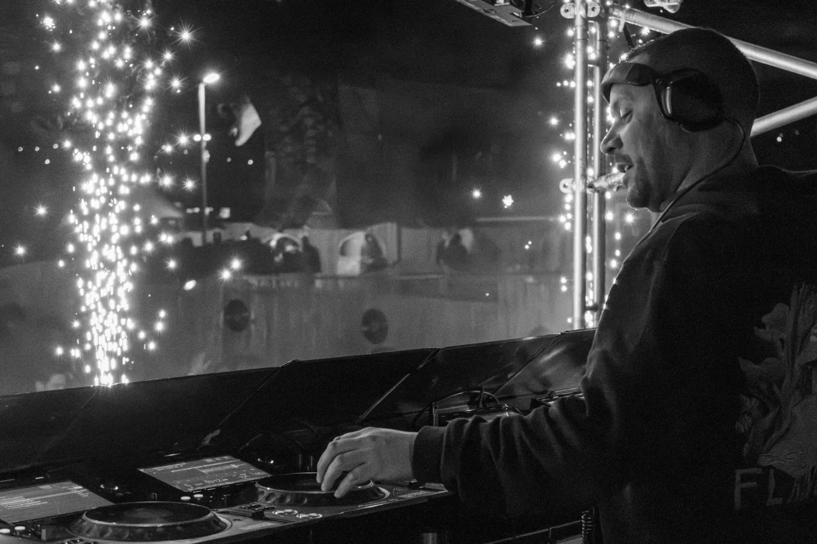 Zwart-wit zijportret van DJ met koptelefoon en sigaar achter draaitafels met vuurwerk fonteinen