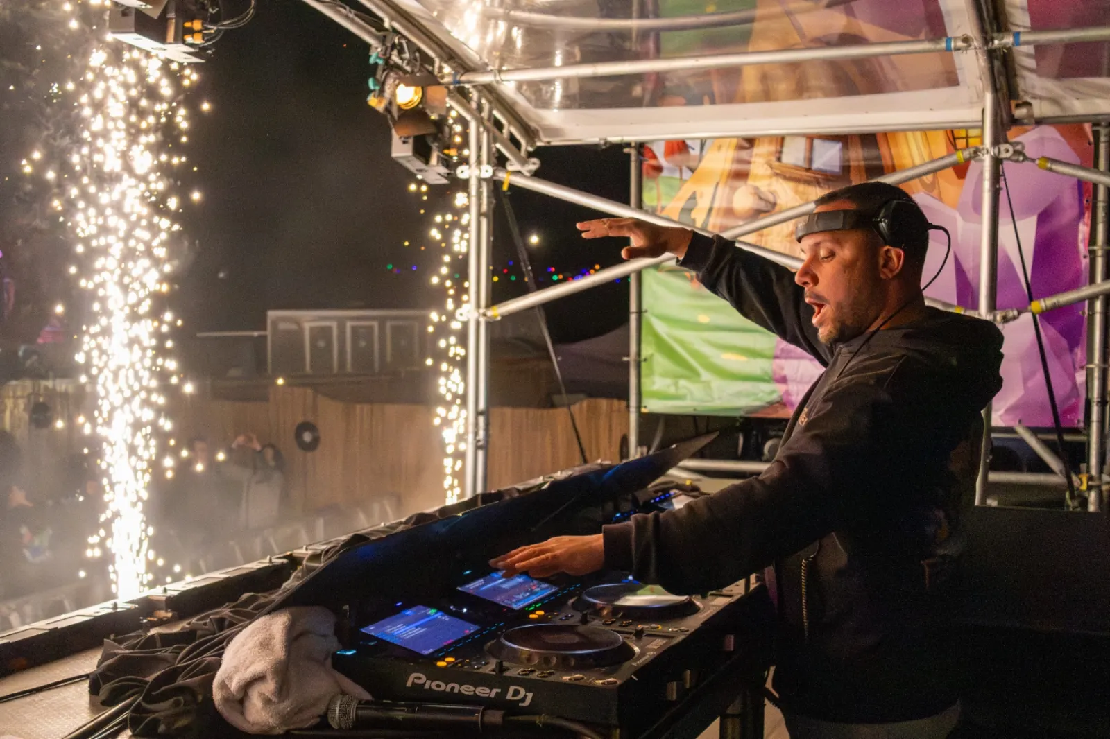 DJ met opgeheven arm achter Pioneer draaitafels naast vuurwerk fonteinen tijdens avondshow