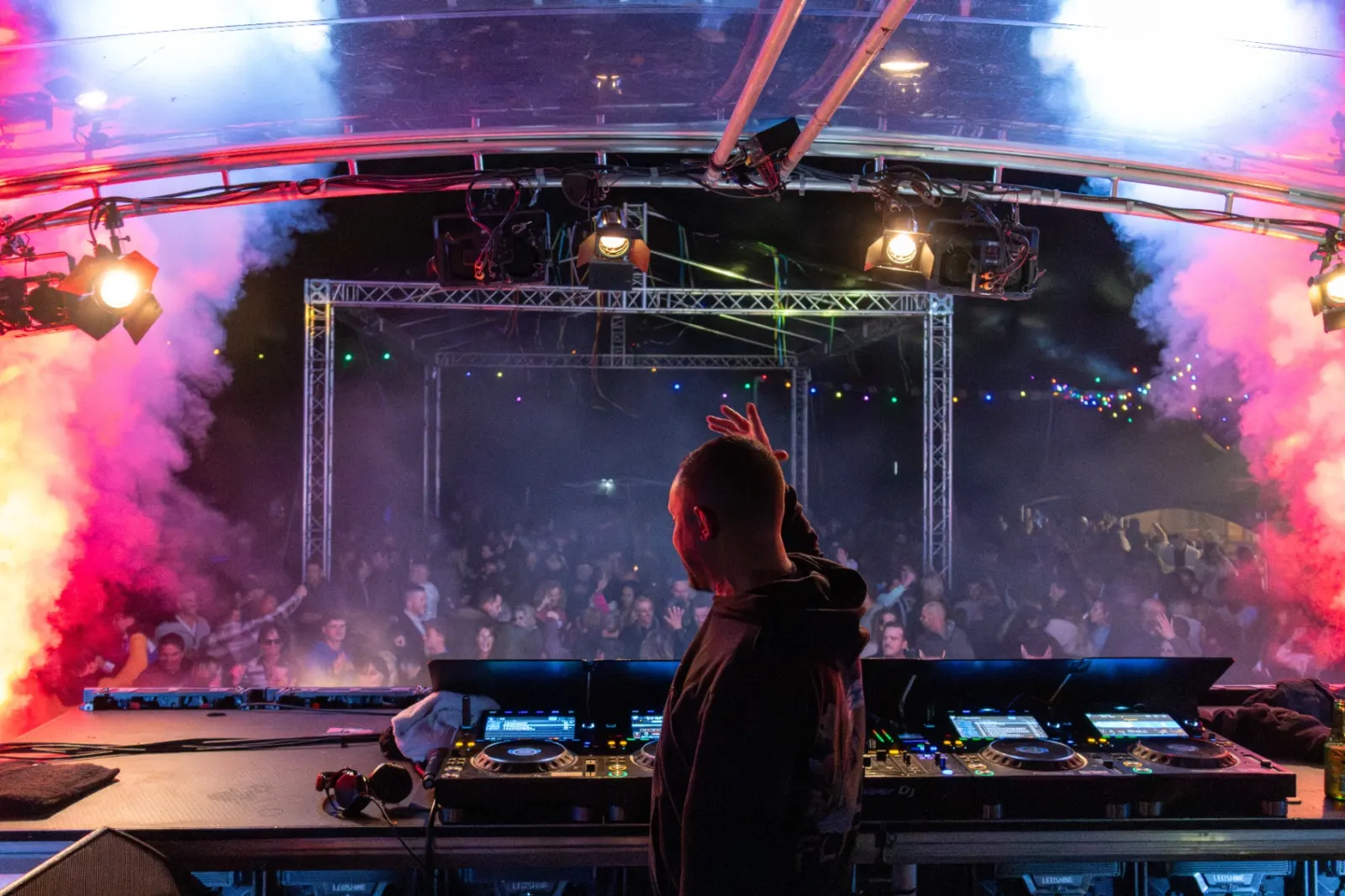 DJ van achteren met opgeheven hand achter Pioneer draaitafels met rode rook en juichend publiek