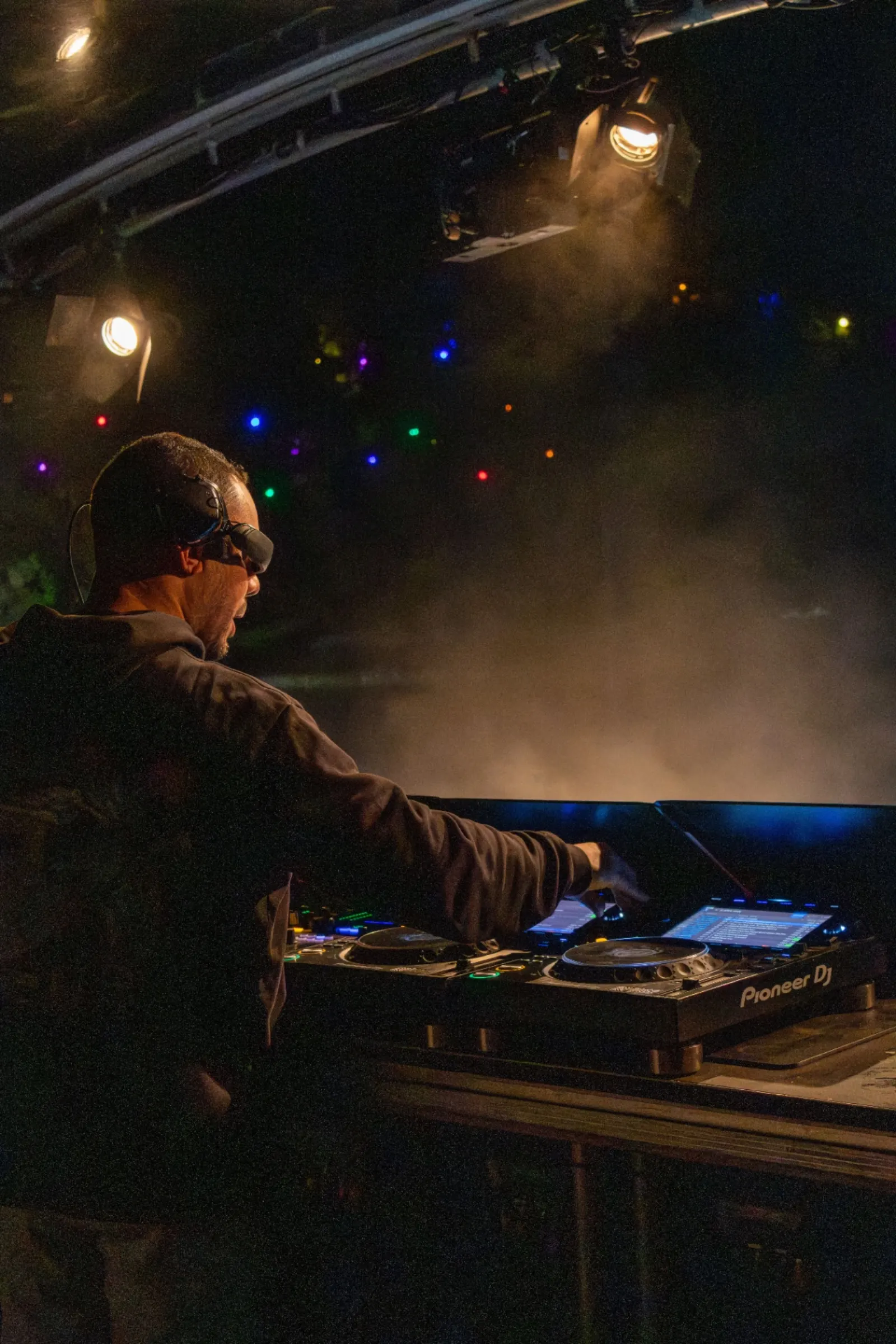 DJ van achteren met koptelefoon en zonnebril achter Pioneer draaitafels onder warm spotlicht in donkere club