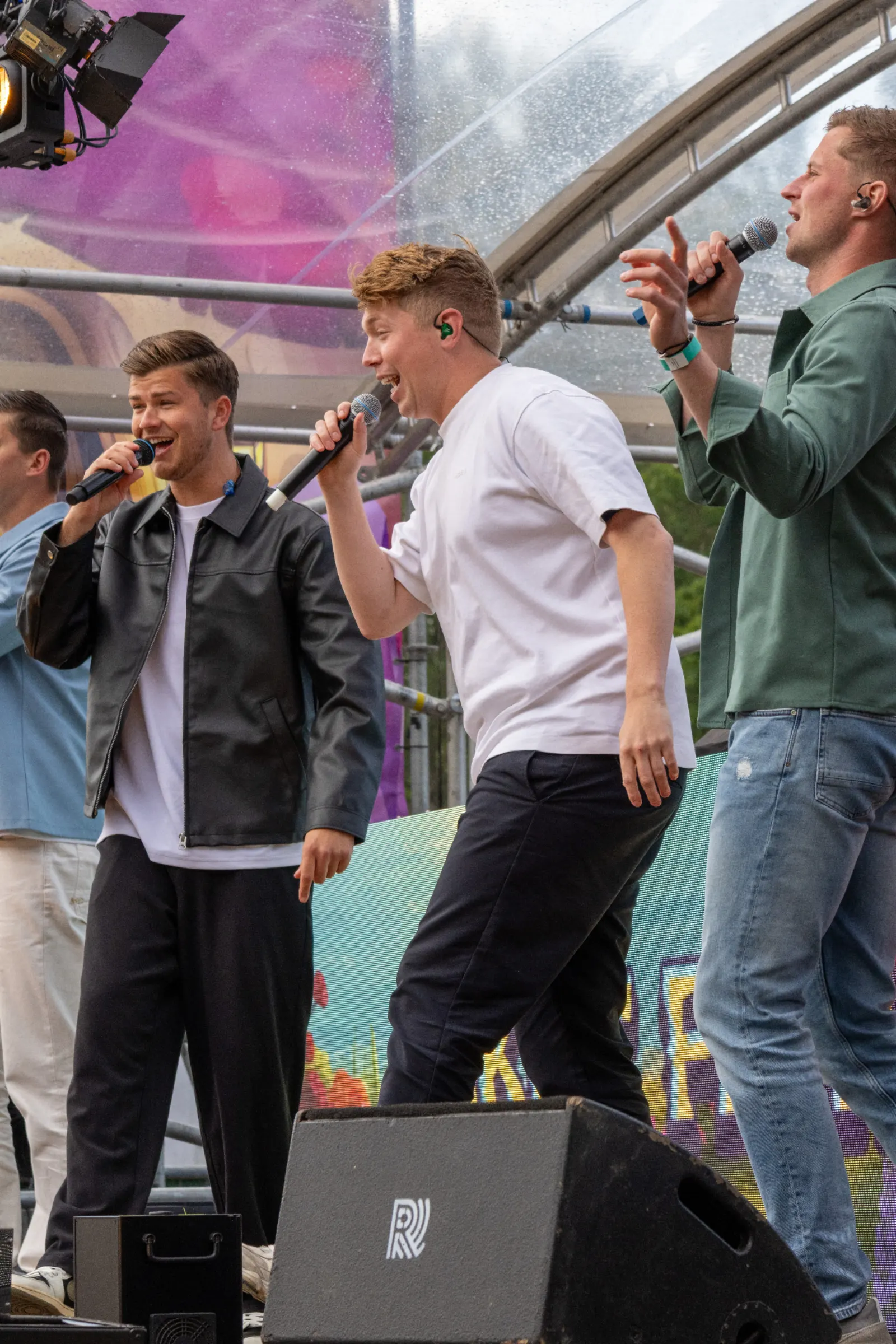 Vier zangers met microfoons op festivalpodium onder transparante koepel met paarse achtergrond