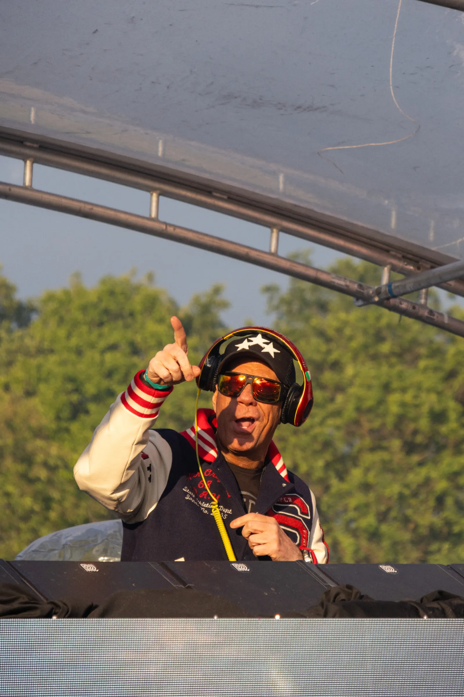 DJ met sterrencap, spiegelende zonnebril en koptelefoon in varsity jack wijst omhoog tijdens openluchtfestival