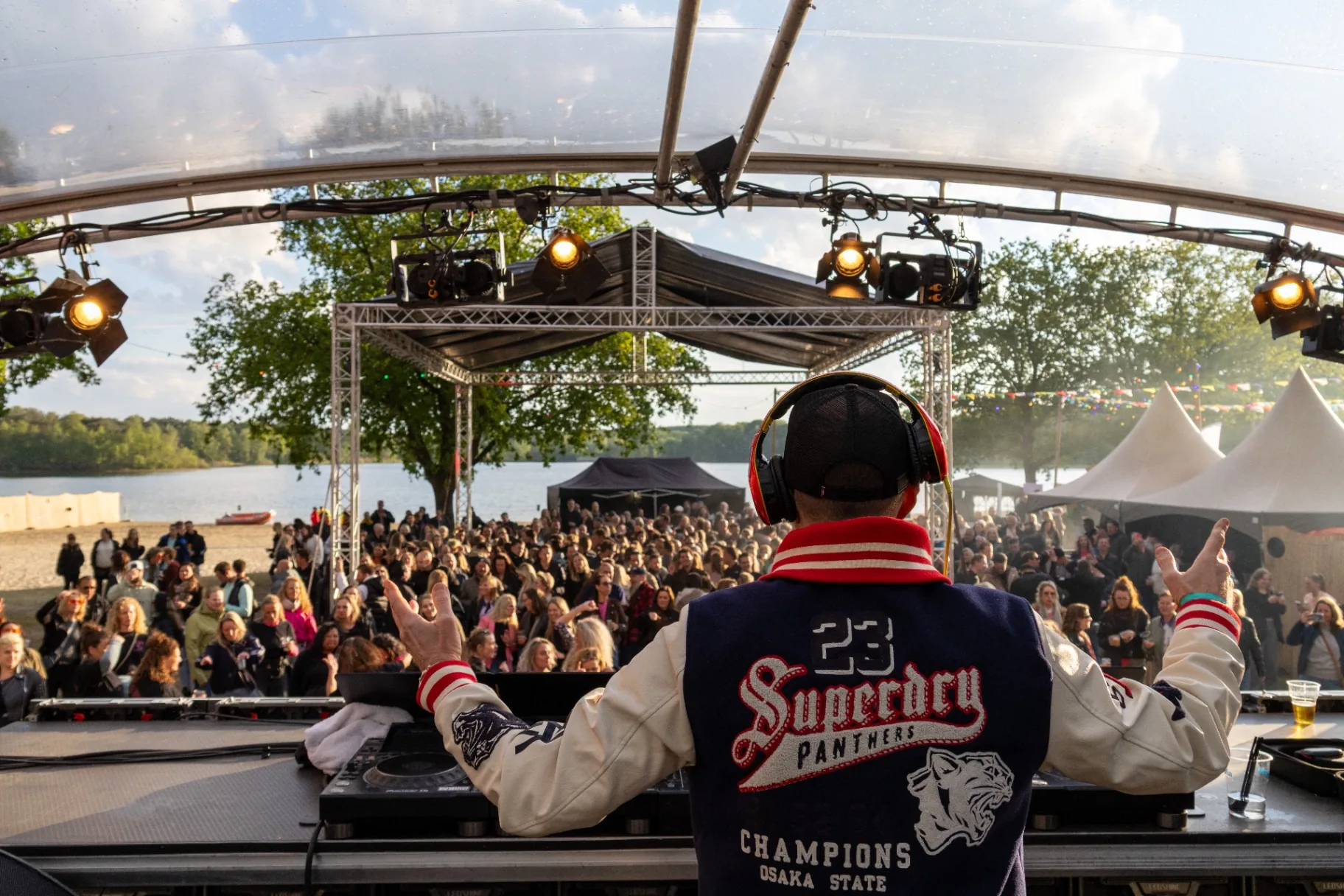 DJ van achteren in varsity jack met koptelefoon en juichend publiek bij zonsondergang aan het meer