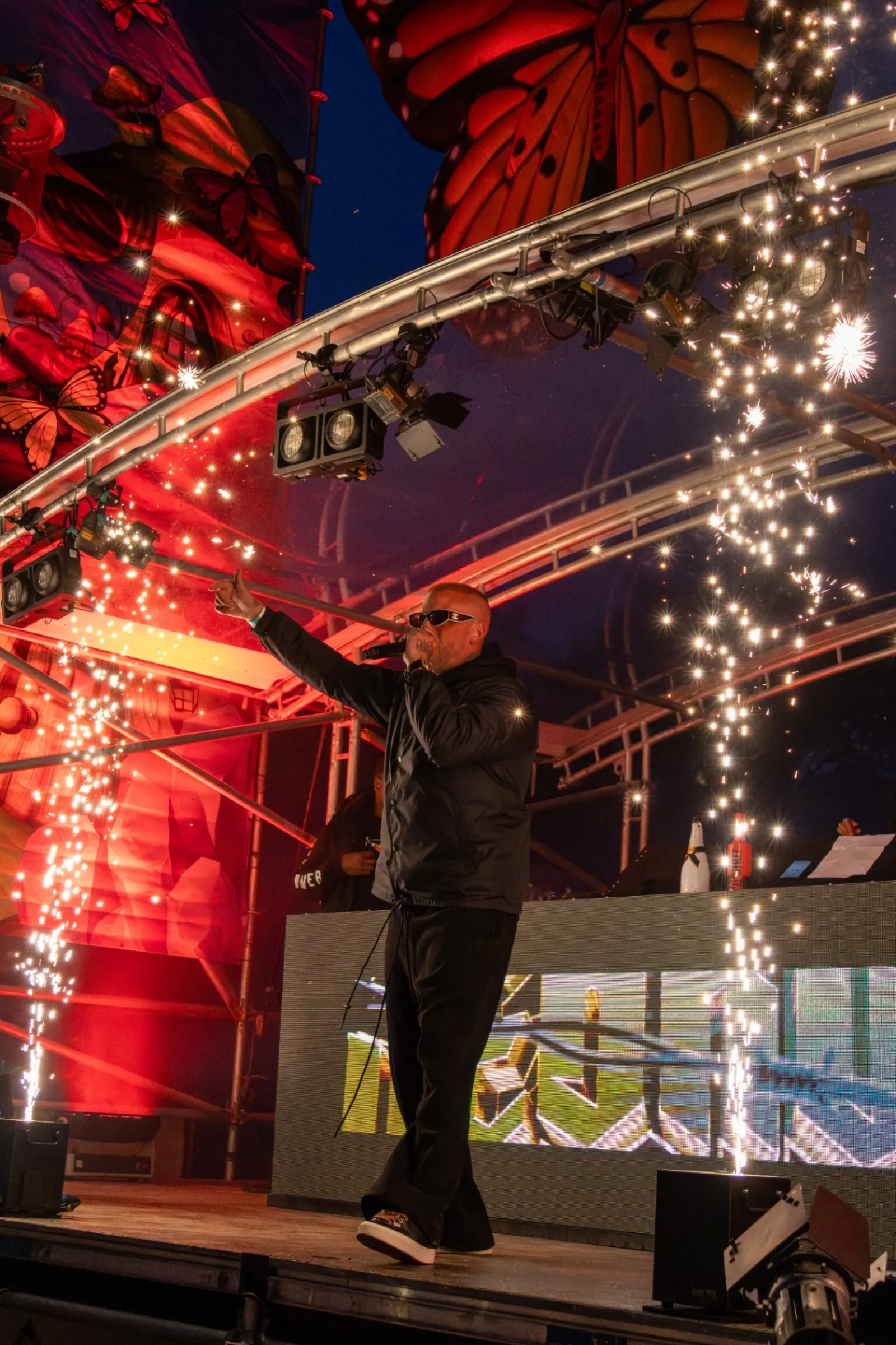 Artiest wijst omhoog tussen vuurwerk fonteinen op festivalpodium