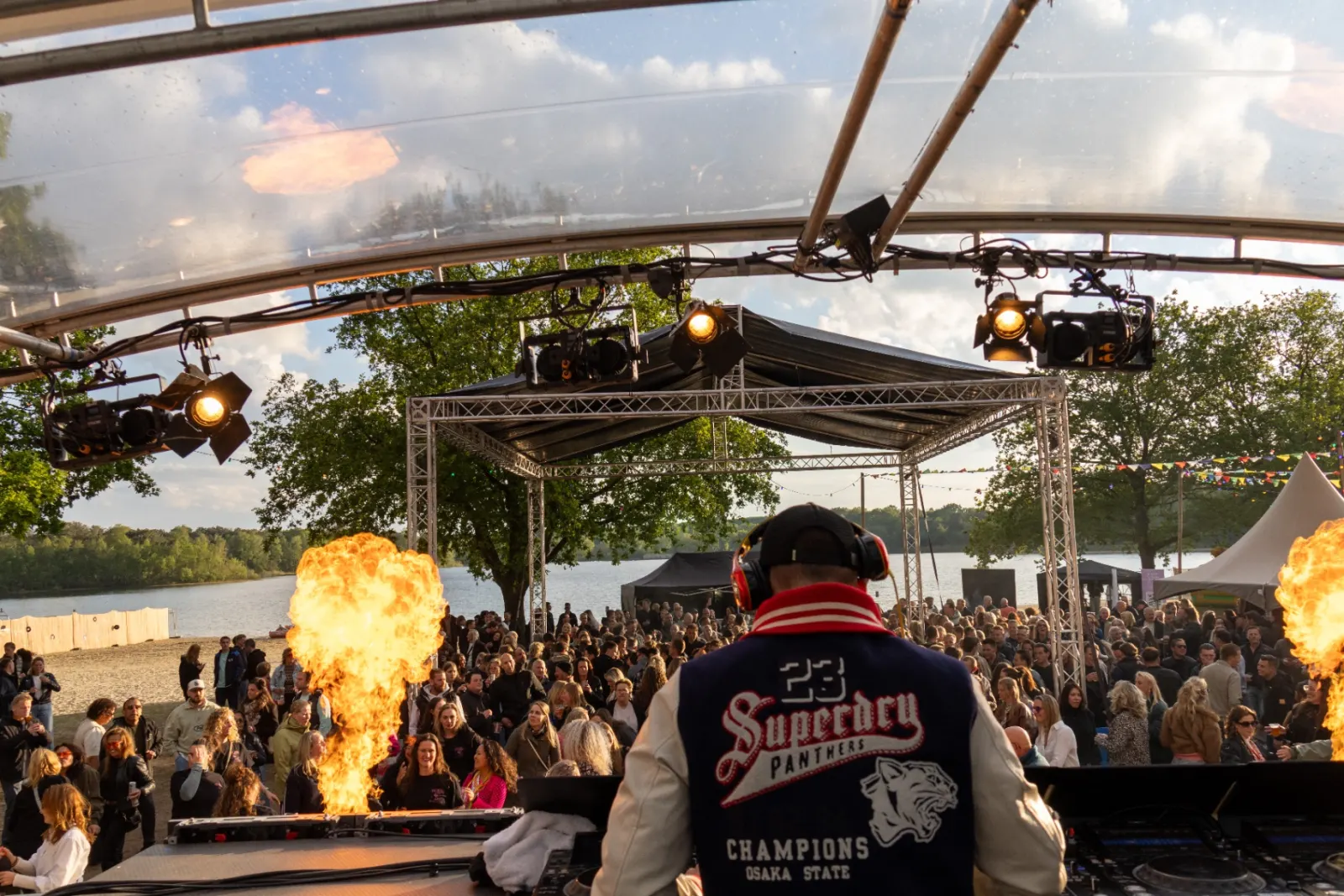 DJ met vuurkanonnen en publiek bij een meer tijdens zonsondergang festival