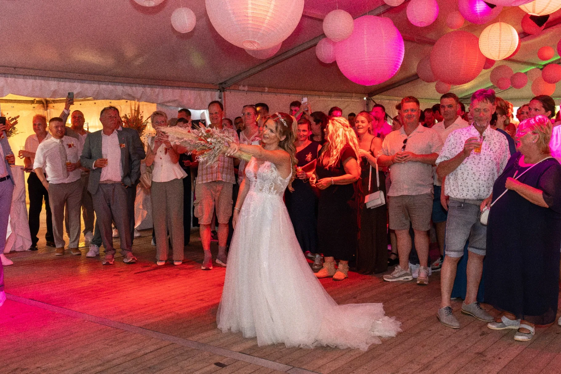 Bruid gooit bruidsboeket onder roze lampionnen met juichende gasten